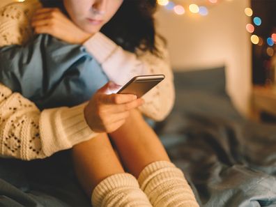 Girl sitting on bed with knees up to their chest using phone.
