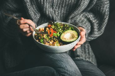Healthy vegetarian dinner. Woman in grey jeans and sweater eating fresh salad, avocado half, grains, beans, roasted vegetables from Buddha bowl. Superfood, clean eating, dieting food concept