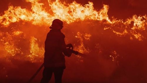 A firefighter stands in the face of a raging fire.