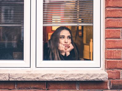 Young woman in lockdown, sitting inside her home, close to the window looking out. She is practicing social distancing due to COVID-19