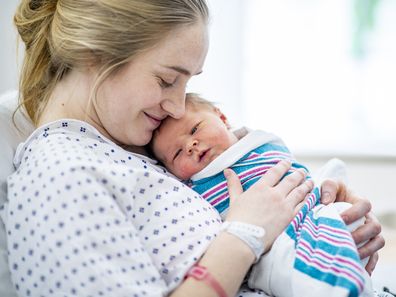 A caucasian mother in a hospital gown sits up in bed and holds her newborn baby to her chest