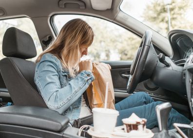 Young woman sitting in her car, opening a take out bag, eating fast food and drinking soda and coffee. In a rush, multi tasking, eating take out burger, french fries and dessert while driving a car.