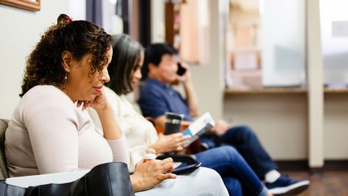 People sit in doctor's waiting room.  One woman looks a medical brochure and a man uses his smart phone to make a call.