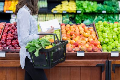 Young woman carries a shopping basket filled with fresh produce. She is shopping for fresh fruit and vegetables in a grocery store.
