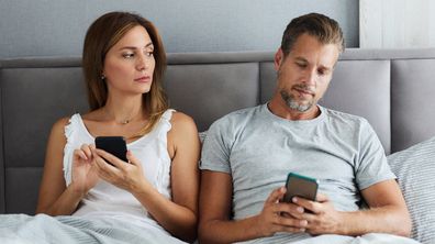 Mid adult couple using mobile phones in a bed while woman is peeking into her husband's phone. cheating affair infidelity