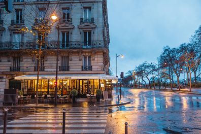 Beautiful traditional French cafe in the corner of a side street in Paris, cafe is closed with chairs on the table, rainy day with wet street in front of the cafe, trees with beautiful early morning sky in the background, horizontal