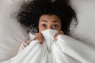Surprised scared funny african girl lying in bed covering face with blanket, young black woman awake from sleep hiding peeking from duvet feel shy afraid or bad dream nightmare embarrassed, top view