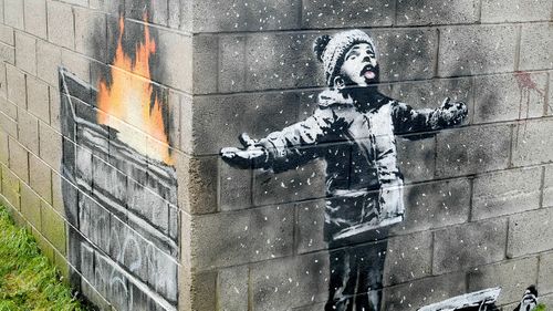 An artwork by Banksy on the side of a garage depicts a child dressed for snow playing in the falling ash and smoke from a skip fire, in Port Talbot, Wales.