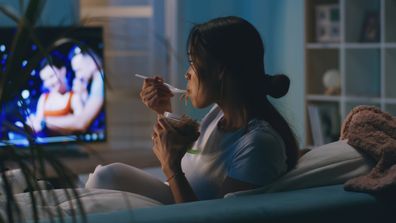 Asian woman eating takeaway noodles and watching TV while sitting on couch and having dinner in evening at home