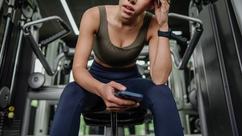 A dedicated young woman seated in the gym, deeply focused on her phone, showcasing the blend of concentration and relaxation during workout routines.
