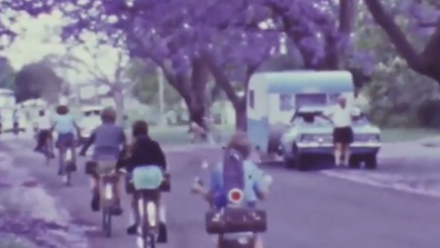 In one shot a group of children are seen riding their bikes as the man poses next to his caravan.