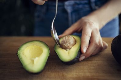 Unrecognizable female person peeling perfectly ripe avocado with spoon on wooden cutting board in kitchen