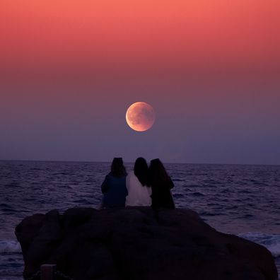 Three women sitting at the shore watching the full moon
