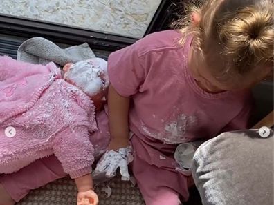 Jen Hawkin's daughter Frankie sitting with her right hand covered in sudocream, holding the container in her left hand, and with her doll on her right with a face covered in sudocream.