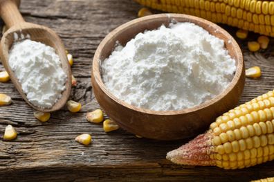 Corn starch, cornflour in bowl and spoon with dried corn groats, kernels on rustic table. Maize starch with ripe cobs and kernels