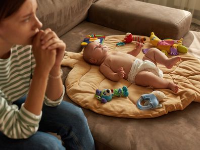 A baby lying on play mat as a sad mum looks away. 