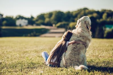 Back view of little cute girl is having fun with golden retriever on a green grass. Charming cheerful girl is sitting with dog labrador.