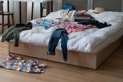modern bright bedroom with messy clothes scatter on white bed and floor. empty room with nobody in cozy apartment. packing luggage suitcase for summer vacation and spring holidays concept lifestyle.