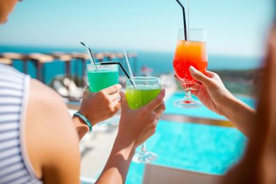Female hands holding glasses of green and orange cocktail, sitting on the terrace in the summer resort and enjoying the moment