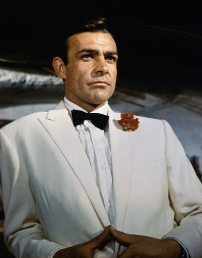 Waist-up portrait of Sean Connery, as James Bond, leaning against a bar and looking out across the room. Connery is wearing a white tuxedo and bow tie with a red carnation in his lapel. CONNERY; SEAN BOND; JAMES. 99/99/1966 35 U CT4X5