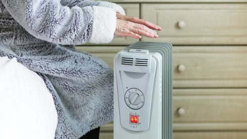 A retired senior woman in her 70s sits at home inside her cold house in winter. It is so cold that she is wrapped up in warm winter clothing, and is holding her hands over an electric heater for some extra warmth and comfort. Selective focus with room for copy space.