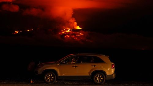 A man talks on a phone in his car alongside Saddle Road as the Mauna Loa volcano erupts Wednesday, Nov. 30, 2022, near Hilo, Hawaii. Hundreds of people in their cars lined Saddle Road, which connects the east and west sides of the island, as lava flowed down the side of Mauna Loa and could be seen fountaining into the air on Wednesday. 