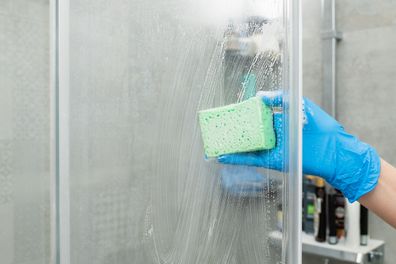 Woman in a blue glove cleans a shower cabin from limestone