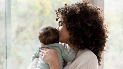 Young mom gives her baby a kiss on his head while standing next to a window in their home