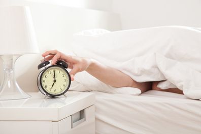 Hand under blanket reaching out for alarm clock, shallow depth of field focus on foreground