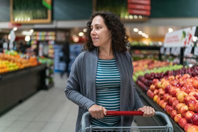 A young mixed race pregnant woman is at the grocery store. She is buying fresh fruit and vegetables in the produce section of the market. The healthy woman is smiling while pushing her grocery cart next to the apples. She is wearing casual maternity clothing.