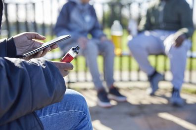 Close Up Of Teenagers With Mobile Phone Vaping and Drinking Alcohol In Park