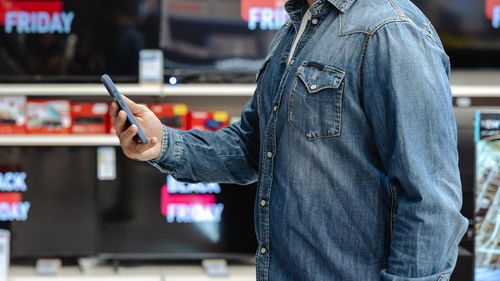 Um jovem procura produtos eletrónicos numa loja durante uma liquidação da Black Friday, segurando o seu smartphone.