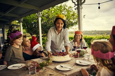 Family Christmas gathering, woman placing pavlova on a table surrounded by other women and children wearing festive paper crowns.
