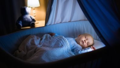 Adorable baby sleeping in blue bassinet with canopy at night. Little boy in pajamas taking a nap in dark room with crib, lamp and toy bear. Bed time for kids. Bedroom and nursery interior.