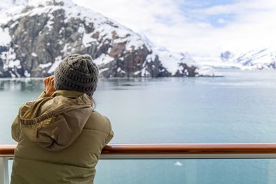 A young woman with binoculars views the snow covered mountains and glaciers from a cruise ship balcony at Glacier Bay National Park and Reserve, Alaska USA.