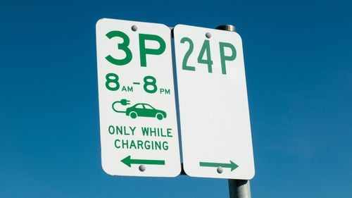 À esquerda está uma placa reservando vagas para veículos elétricos e limites de tempo para recarga. A placa à direita indica tempo de estacionamento ilimitado de 24 horas. Esta imagem foi tirada em um estacionamento público próximo ao campo esportivo de Heffron, Maroubra, Sydney, em uma tarde ensolarada de 13 de abril de 2025.