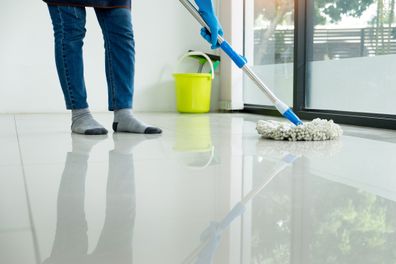 Young housekeeper cleaning floor mobbing holding mop and plastic bucket with brushes, gloves and detergents in the leaving room house floor helping his wife