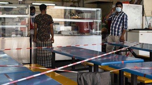 Migrant workers wearing protective face masks walk past tables and chairs in a canteen with barricade.