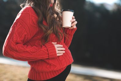 Stylish pregnant woman drinking coffee outdoors wearing red knitted sweater outdoors closeup. Motherhood. Maternity.