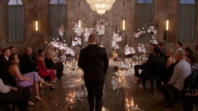 A groom walks down down the aisle to await his bride. There are candles on the floor and an oversized chandelier overhead, creating spectacular, opulent lighting.