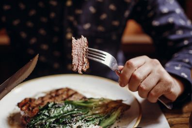 Hands holding fork and knife and eating delicious juicy steak with grilled cabbage,tomatoes and cheese on table at cafe in city street. 