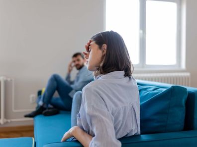 Stressed woman in the foreground sitting on a couch with her hand in her head, he partner sits on the end of the couch looking at her with his hand on his head.