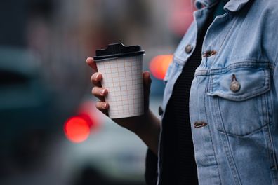 Close up of an unrecognizable woman in denim jacket, walking outside and holding a paper coffee cup to go.