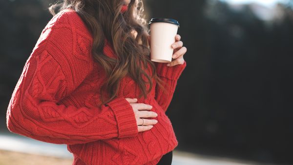 Stylish pregnant woman drinking coffee outdoors wearing red knitted sweater outdoors closeup. Motherhood. Maternity.