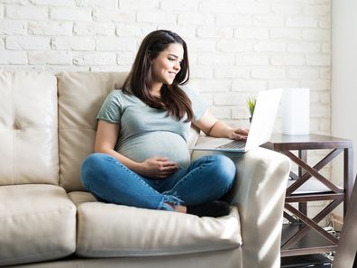 Full length of Caucasian pregnant female using laptop while sitting on sofa in living room
