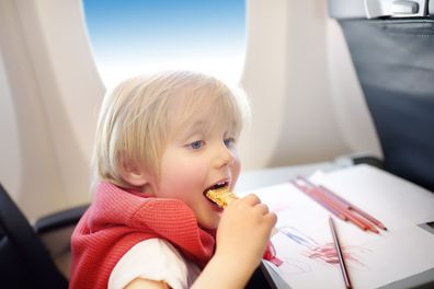 Charming kid traveling by an airplane. Joyful little boy sitting by aircraft window during the flight. Child drawing picture and eating snack. Air travel with little kids