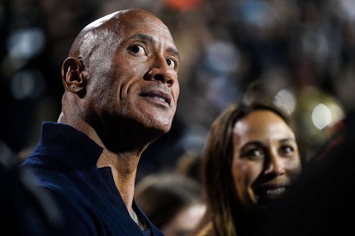 Dwayne 'The Rock' Johnson stands on the sideline in a football game.