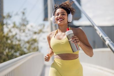 Portrait of a cheerful woman with wireless headphones running on a sunny day. She is holding a water bottle while running.