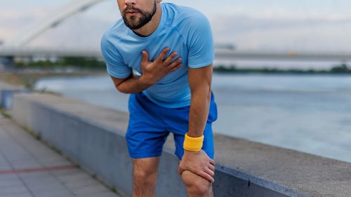 Um homem de camisa azul claro e short azul se exercita ao ar livre à beira do rio, segurando o peito e curvado, com uma ponte ao fundo, transmitindo cansaço.