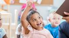 Pretty preschool age girl raises her hand to ask or answer a question in her classroom. An unrecognizable teacher is reading the class a story.